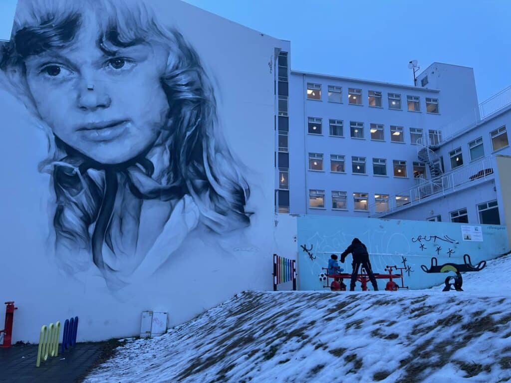 kinder-spielen-auf-spielplatz-im-akureyri-vor-gebaeude-mit-grossem-streetartbild-eines-maedchengesichts-in-der-daemmerung