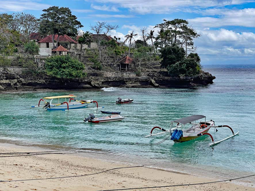 sandstrand-mit-booten-und-blauem-wasser-am-rand-felskueste-mit-haeusern-nusa-lembongan