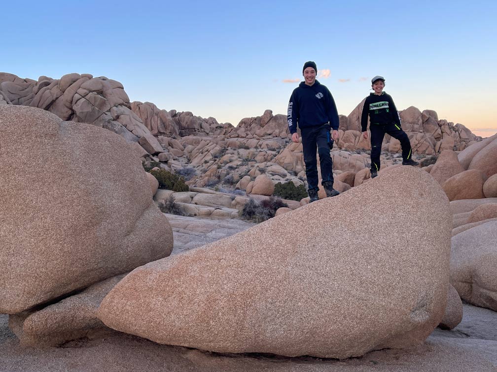 zwei-kinder-stehen-auf-felsen-im-joshua-tree-nationalpark-rundherum-weitere-felsen-im-rosa-sonnenuntergang-himmel-