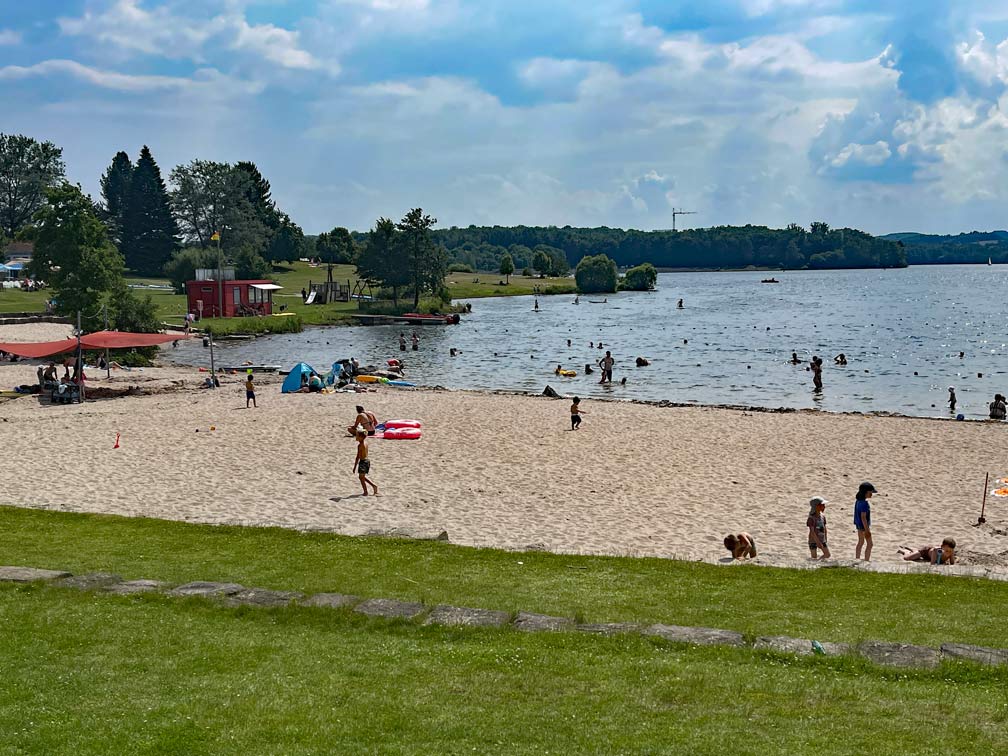 gruene-wiese-grenzt-an-sandstrand-am-badesee-bostalsee-spielende-kinder-sind-zu-sehen