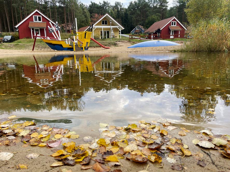 kleiner-badesee-im-herbst-mit-bunten-blaettern-dahinter-spielplatz-und-bunte-ferienhaeuser